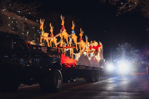 A festive parade float featuring reindeer and Santa, illuminated at night, with a truck in the foreground.