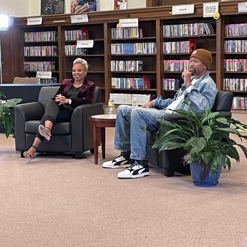 Two speakers sit in armchairs during a discussion, surrounded by plants and bookshelves in a cozy setting.