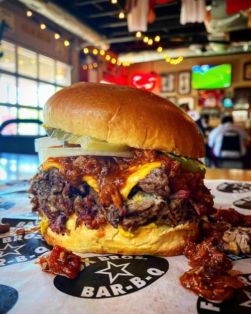 A close-up of a juicy burger with melted cheese, pulled meat, pickles, and barbecue sauce on a bun, served in a restaurant.