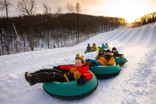 A group of children enjoying snow tubing down a snowy hill at sunset, wearing colorful winter jackets.