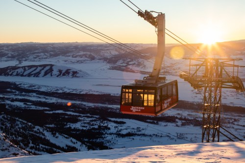 A ski gondola glides over a snowy landscape at sunset, with mountains and valleys in the background.