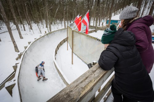 A person sleds down a snowy track while two spectators watch from a wooden railing above. Flags are visible in the background.