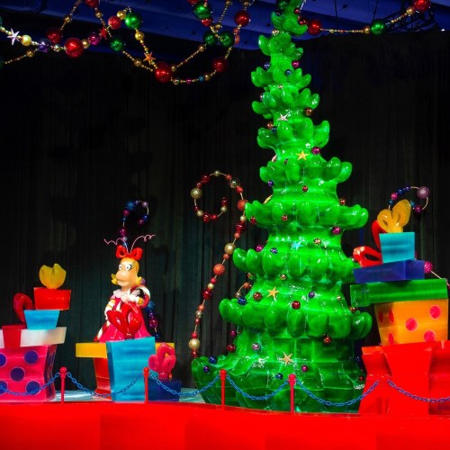 A colorful, festive display featuring a large green Christmas tree and wrapped gifts, with a whimsical character nearby.