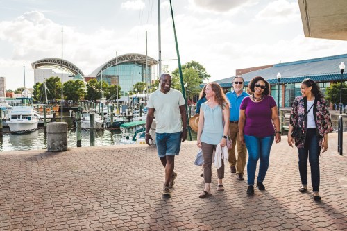 A diverse group of five people walks along a waterfront promenade near boats and buildings, enjoying a sunny day.