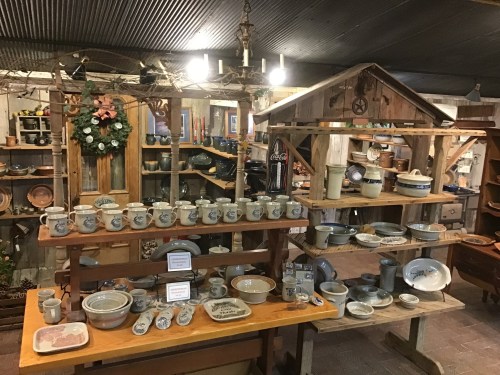A rustic shop display featuring various pottery and ceramic items on wooden shelves in a cozy, decorated space.