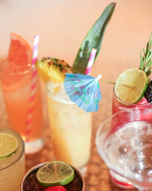 A colorful assortment of cocktails with garnishes, including fruit, umbrellas, and straws, on a textured surface.