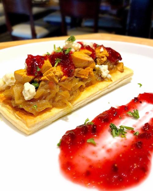 A flaky pastry topped with chicken, caramelized onions, and a red berry sauce, garnished with herbs.