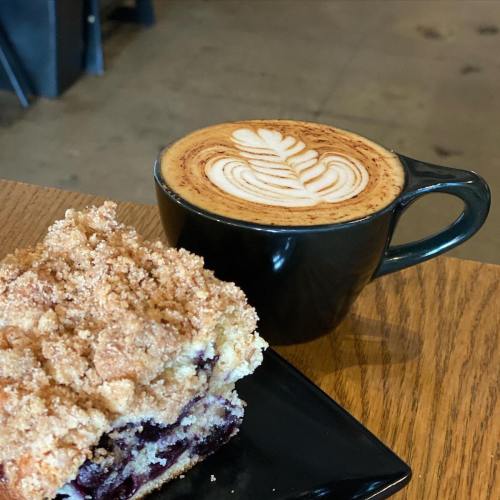 A slice of blueberry coffee cake next to a latte with a leaf design, on a wooden table.