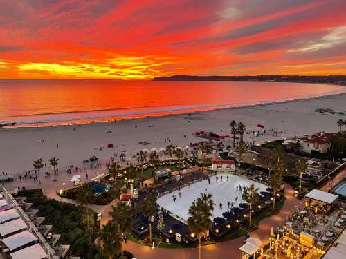 A vibrant sunset over a beach with an ice skating rink, palm trees, and people enjoying the view.
