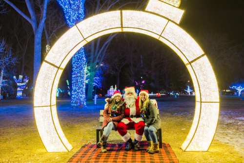 A festive scene with Santa sitting on a bench, flanked by two smiling women, surrounded by colorful holiday lights.