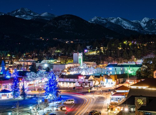 A scenic night view of a mountain town adorned with colorful lights, surrounded by snow-capped peaks.