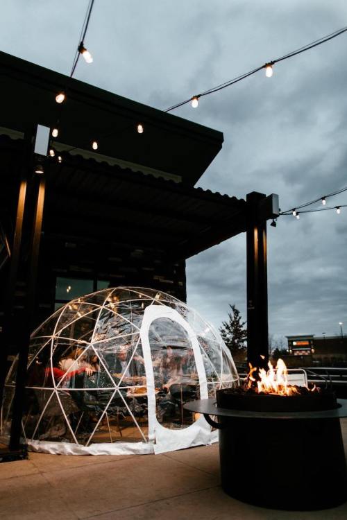 A clear igloo-style dining pod next to a fire pit, with string lights overhead and a cloudy sky in the background.