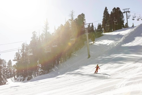 A skier glides down a snowy slope with ski lifts and trees in the background, illuminated by bright sunlight.