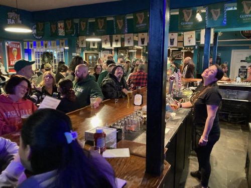 A lively bar scene with a crowd enjoying drinks, a bartender serving, and festive decorations in the background.