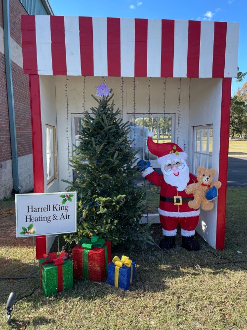 A festive display featuring a decorated Christmas tree, Santa holding a teddy bear, and colorful gift boxes outside a booth.