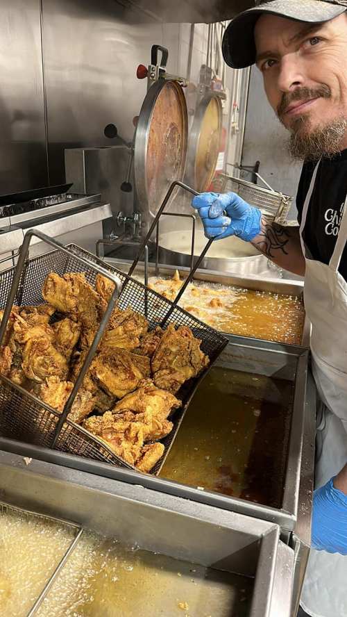 A chef in a kitchen holds a basket of golden fried chicken over a fryer, with oil bubbling in the background.
