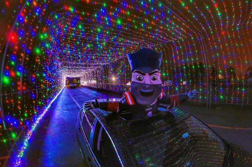 A mascot with a big smile leans out of a car, surrounded by a colorful tunnel of holiday lights.