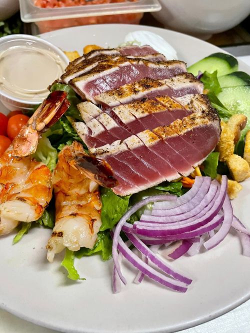A colorful salad with grilled tuna, shrimp, cucumbers, red onion, and a small cup of dressing on the side.