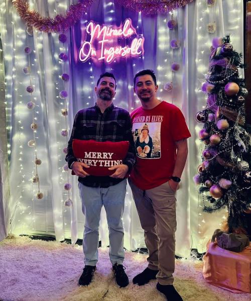 Two men pose together in front of a festive backdrop with lights and a Christmas tree, holding a "Merry Everything" pillow.