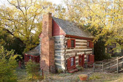 A rustic log cabin with red shutters, surrounded by trees in autumn foliage, showcasing a historic charm.