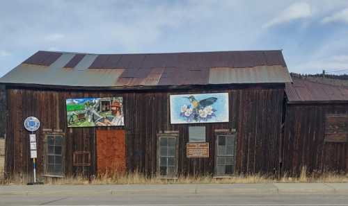 Weathered wooden building with murals of a landscape and a hummingbird, featuring a rusty metal roof.