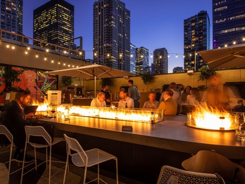 A rooftop bar at dusk with a glowing fire feature, city skyline, and people socializing in a vibrant atmosphere.