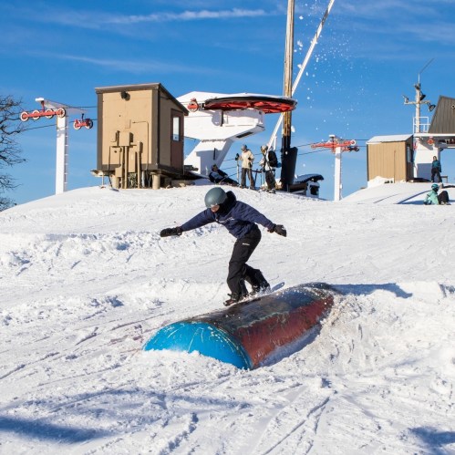 A snowboarder performs tricks on a rail in a snowy terrain park under a clear blue sky.