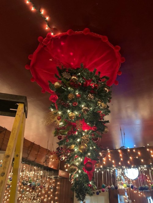 A festive upside-down Christmas tree adorned with ornaments, hanging from a ceiling decorated with lights.