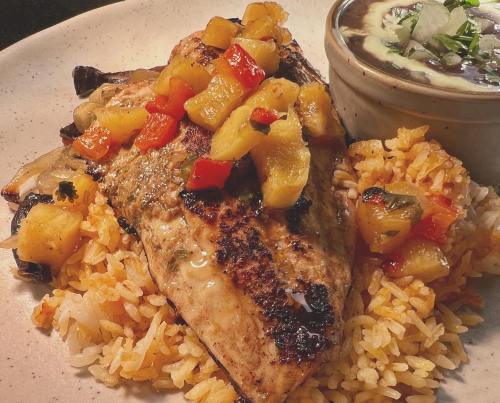 Grilled fish topped with pineapple salsa, served on a bed of rice with a side of black beans and onions.