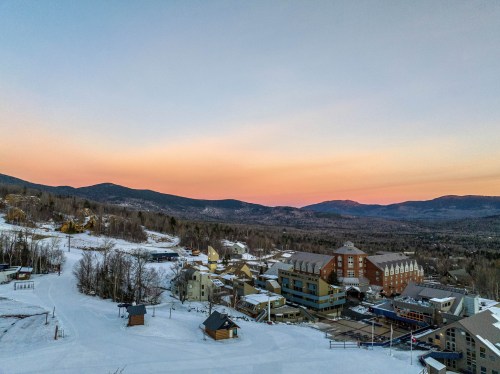 A scenic view of a snowy landscape at sunset, featuring mountains and a ski resort with buildings below.
