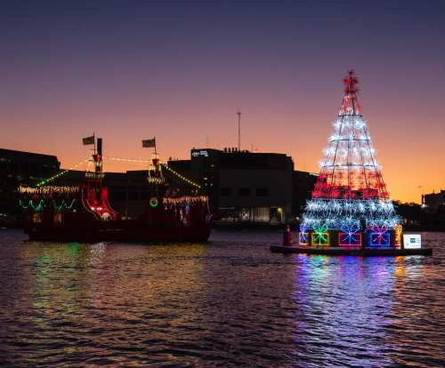 A brightly lit boat and a colorful Christmas tree float on water at sunset, creating a festive scene.