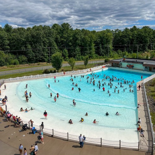 A large outdoor pool filled with people enjoying a sunny day, surrounded by trees and a cloudy sky.