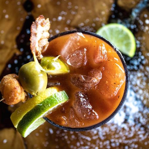 A close-up of a cocktail with ice, garnished with shrimp, olives, lime slices, and a sprinkle of salt.