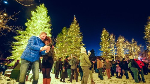 A festive crowd gathers around lit Christmas trees, with a couple taking a selfie in the foreground.