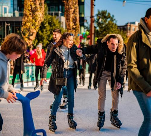A couple enjoys ice skating, laughing as they hold hands on a busy rink surrounded by other skaters.