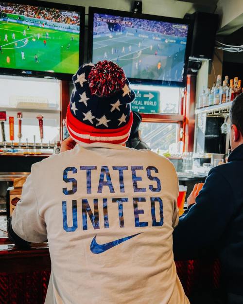 A person wearing a "States United" shirt watches a soccer game on multiple screens in a bar.