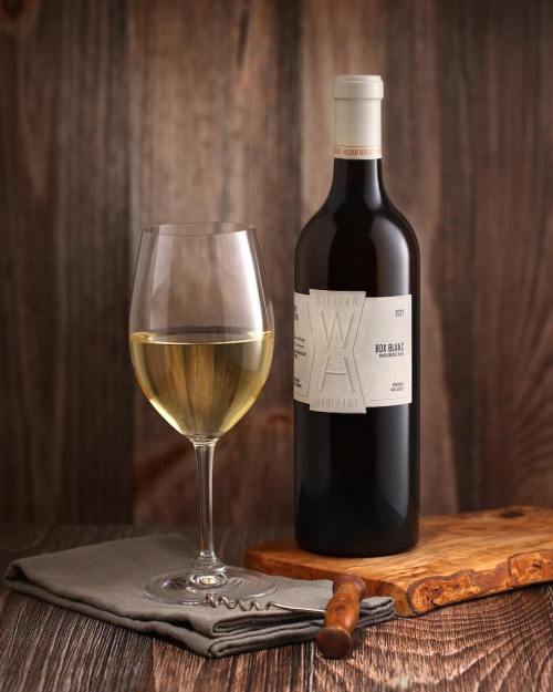 A bottle of white wine next to a glass of wine on a wooden surface, with a cloth napkin and a corkscrew.