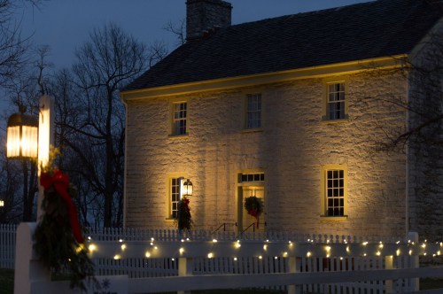 A historic stone house illuminated at dusk, decorated with wreaths and string lights, surrounded by a white picket fence.