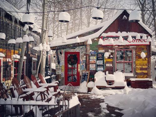 A colorful, rustic coffee shop covered in snow, with rocking chairs and hanging lights in a winter setting.