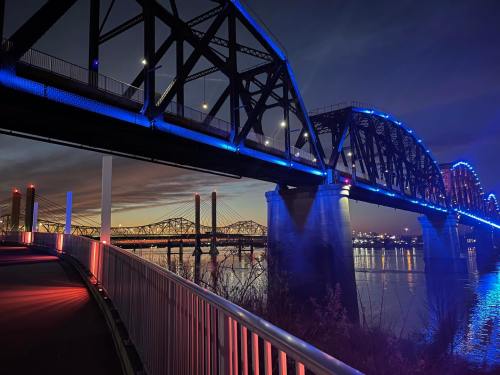 A illuminated bridge at night, reflecting on the water, with colorful lights and a scenic skyline in the background.