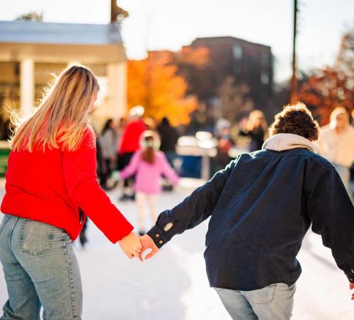 A couple ice skating, holding hands, with people and colorful autumn trees in the background.
