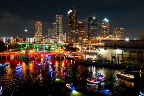 A vibrant night scene of a city skyline with colorful boats illuminated on the water, reflecting city lights.