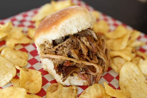A pulled pork sandwich on a bun, surrounded by crispy potato chips on a red and white checkered paper.