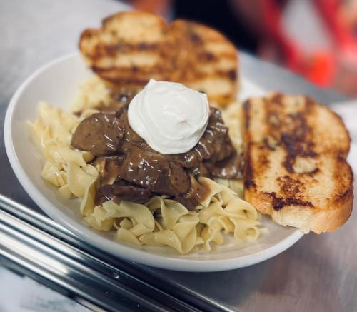 A plate of egg noodles topped with beef and gravy, accompanied by two slices of toasted bread and a dollop of sour cream.
