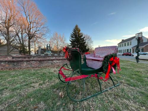 A festive sleigh decorated with ribbons sits on green grass, with trees and buildings in the background under a clear sky.
