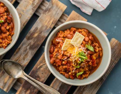 A bowl of chili topped with cheese, crackers, and herbs, served on a wooden surface with a spoon nearby.