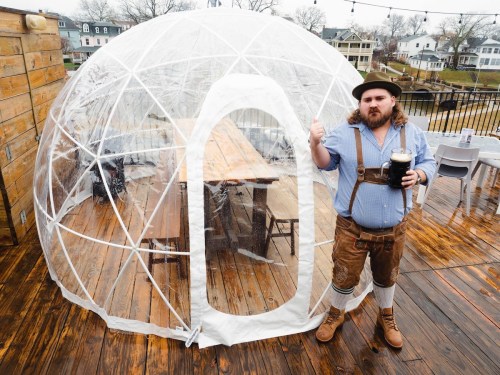 A man in traditional attire stands next to a clear igloo-like structure on a wooden deck, holding a drink.