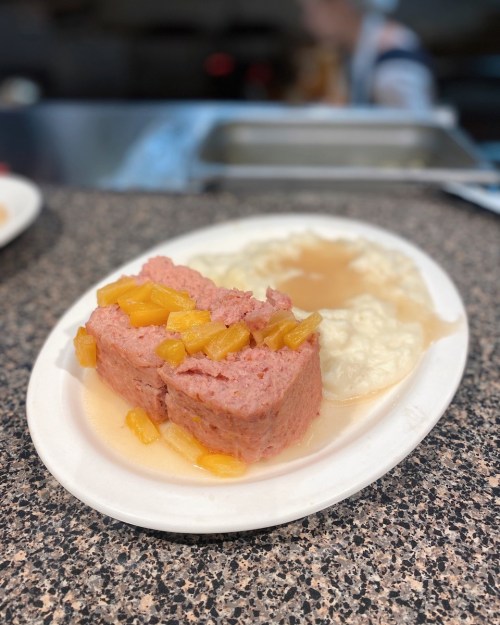 A plate with sliced meatloaf topped with pineapple, served alongside a portion of mashed potatoes.