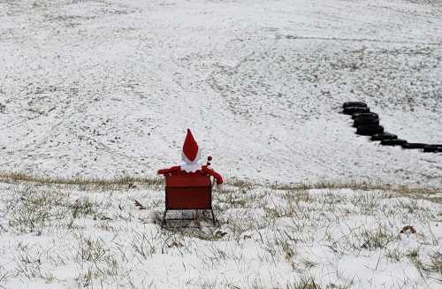 A small red sleigh with a Santa figure sits on a snowy hillside, surrounded by grass and a tire pile in the background.