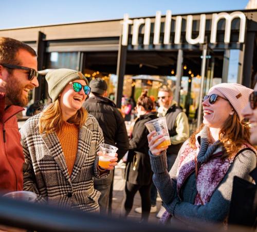 A group of friends laughing and enjoying drinks outdoors at Trillium, with a lively crowd in the background.
